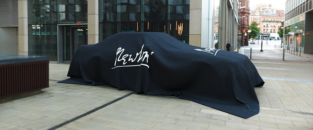 Автомобиль Newton под чёрным покрывалом с автографом у стеклянного фасада в городе