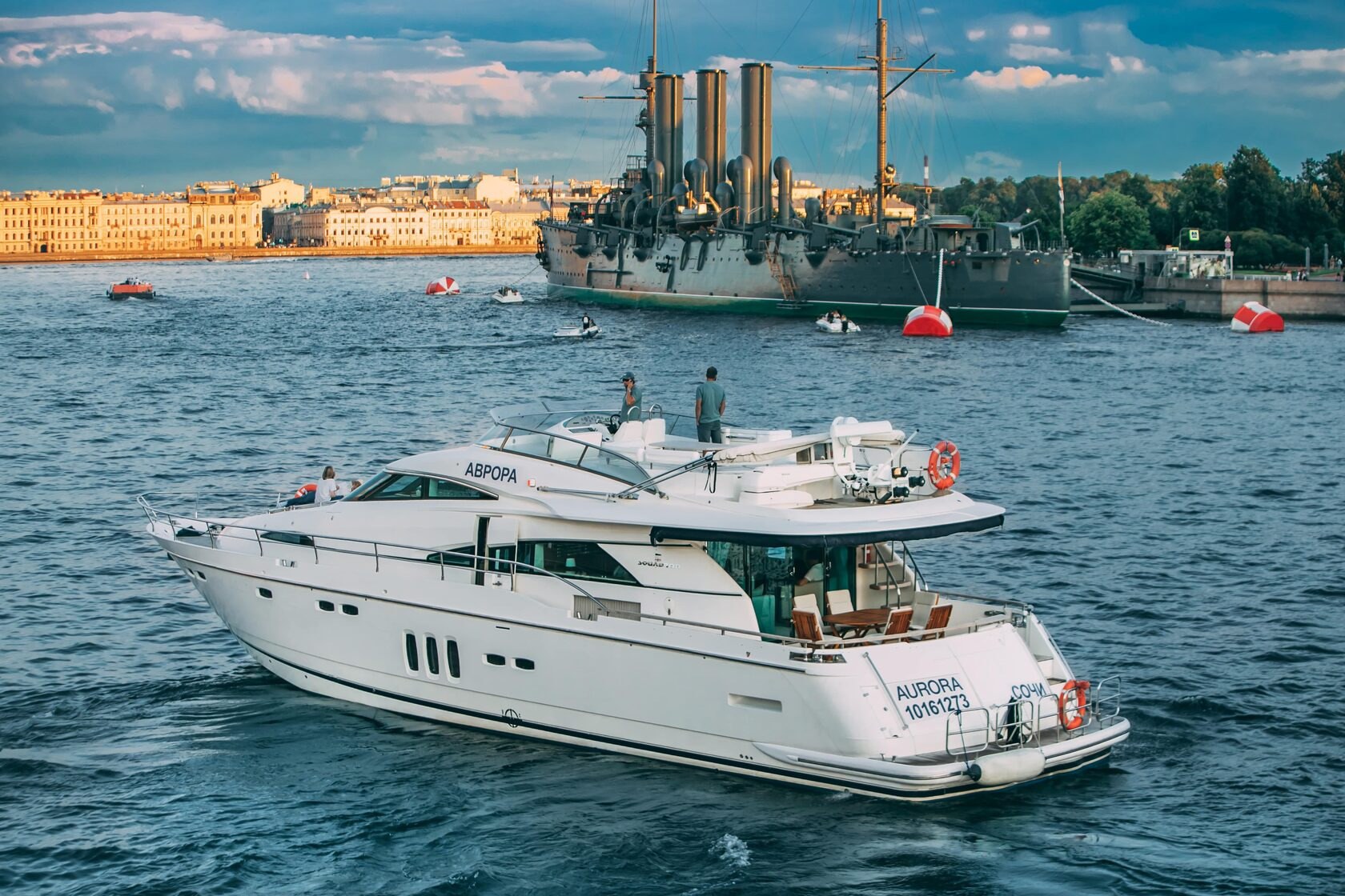Белая моторная яхта Fairline 68 «Аврора» на воде в Петербурге, за ней крейсер «Аврора» и дома на набережной